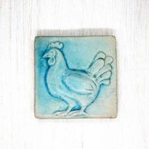 Könnte beinhalten: Eine blaue Keramikfliese mit einem erhabenen Design eines weißen Huhns. Das Huhn ist zur rechten Seite der Fliese gewandt.