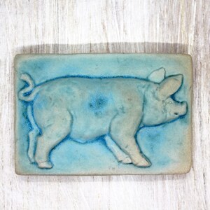 Könnte beinhalten: Rechteckige Keramikfliese mit hellblauem Hintergrund und einem Relief eines Schweins. Das Schwein ist hellgrau mit dunkelblauen Akzenten, auf einer weißen Holzoberfläche.