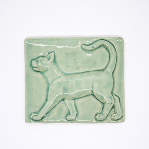 Könnte beinhalten: Eine rechteckige Keramikfliese mit hellminzgrüner Glasur und einem erhabenen Reliefdesign einer Katze im Profil. Die Katze ist gehend dargestellt, wobei ihr Schwanz nach oben gebogen ist, was eine glatte, glänzende Oberfläche aufweist.