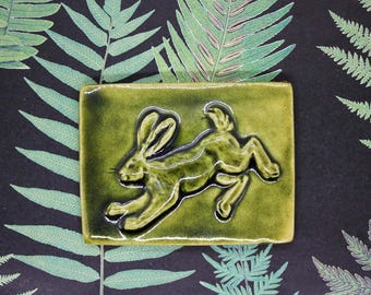 May include: A green ceramic tile with a raised image of a rabbit in a running pose. The rabbit is in a light brown color and is set against a dark green background.