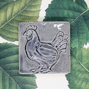 Puede incluir: Un azulejo de cerámica gris con un diseño en relieve de un pollo. El pollo está mirando hacia la izquierda y tiene un diseño detallado de plumas.