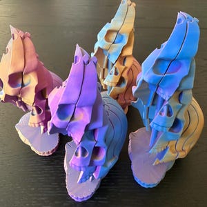 May include: Four 3D-printed articulated skull sculptures featuring a gradient color scheme of purple, blue, and orange. Each figure consists of stacked, segmented skull components that create a vertical, flowing structure with a textured base.