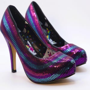 May include: A pair of high-heeled shoes with a sequined design in shades of purple, blue, and pink. The shoes have a pointed toe and a stiletto heel. The interior lining features a whimsical pattern. The heels measure approximately 10 cm.