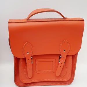 Peut inclure: Sacoche en cuir orange vif avec une poignée supérieure et une fermeture à rabat sécurisée par deux sangles à boucles. Le sac a une poche avant rectangulaire. Idéal pour les livres.