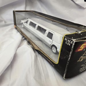 Może przedstawiać: Biały model zabawkowy limuzyny Lincoln w czarnym pudełku. Model w skali 1:24. Na pudełku widnieje napis "Lincoln Limousine". Zabawkowy samochód ma srebrne koła i białe nadwozie.