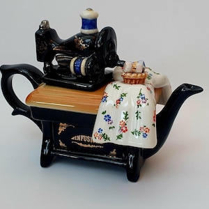 Può includere: Teiera in ceramica nera e oro a forma di macchina da cucire vintage. La teiera presenta una superficie effetto legno, una bobina blu e bianca e un grembiule a fantasia floreale. La teiera ha un manico e un beccuccio.