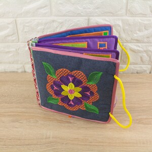 Puede incluir: Un libro suave, similar al denim, con una flor bordada de colores en la portada. El libro tiene un asa de cordón amarillo y está hecho de varias capas de tela.