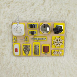 Puede incluir: Un tablero de actividades de madera con varios elementos interactivos para el juego sensorial. Incluye un interruptor de luz, pestillo, espejo, engranajes y un timón. El tablero tiene un acabado de madera natural con detalles amarillos.