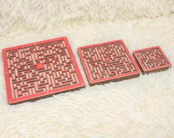 May include: Three wooden maze puzzles of varying sizes. The mazes are red and brown with a white background. The mazes are on a white fluffy surface.