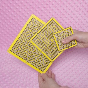 Hands holding three yellow plywood labyrinth mazes in small, medium, and large sizes against a soft pink background, showcasing a handmade maze game set.