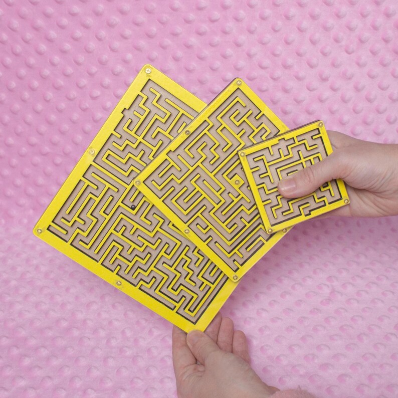 Hands holding three yellow plywood labyrinth mazes in small, medium, and large sizes against a soft pink background, showcasing a handmade maze game set.