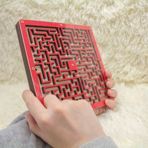 Backside view of a double-sided wooden labyrinth maze held in hands. The red maze design is visible, crafted with layered wood, shown against a fluffy cream background. Even double-sided, have only one metal bead.