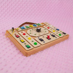 May include: A wooden sensory board featuring various buttons, switches, and a dial. The name "MASON" is displayed in colourful letters. The board includes a patterned handle. The background is a soft pink fabric.