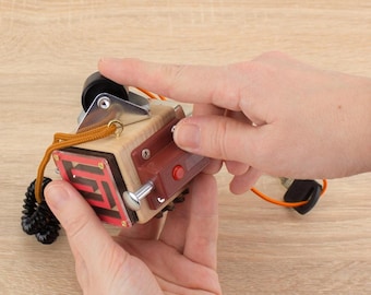 May include: A wooden puzzle box with a red button, a metal lever, and a maze design on the front. The box is being held in a person's hands.