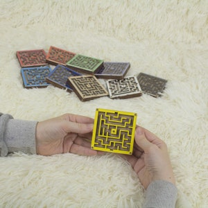 Hands holding a small wooden labyrinth yellow maze with a metal bead, in front of a colorful assortment of similar maze maze in various colors like red, blue, green, yellow, and more. All displayed on a soft cream fur background.