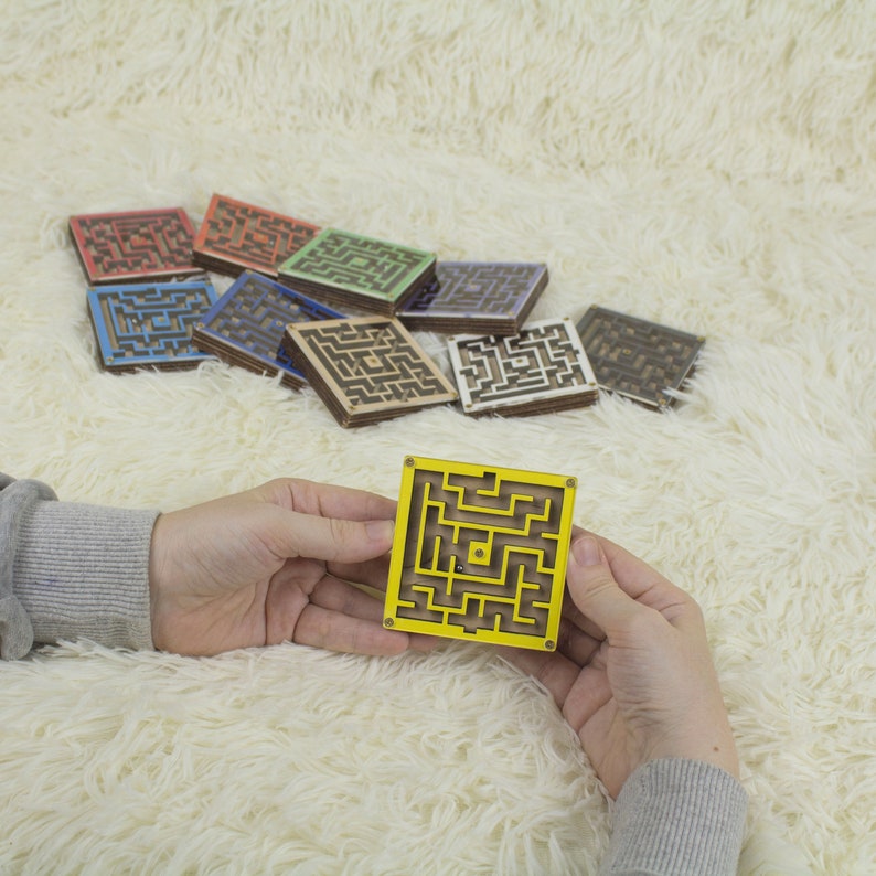 Hands holding a small wooden labyrinth yellow maze with a metal bead, in front of a colorful assortment of similar maze maze in various colors like red, blue, green, yellow, and more. All displayed on a soft cream fur background.