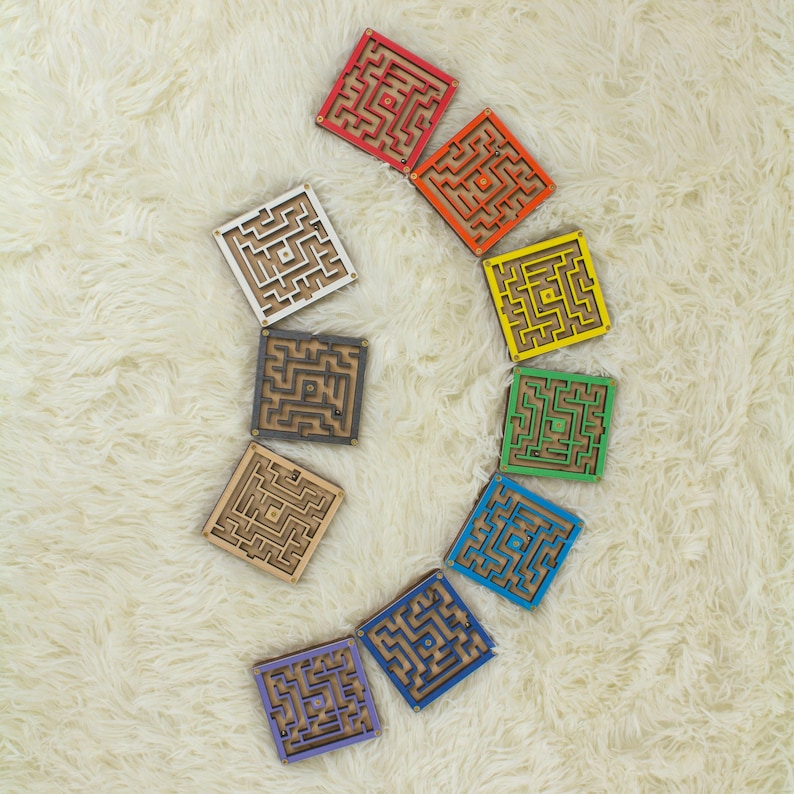 Ten small wooden marble maze toys displayed on a soft cream background, each labeled with a different color option: Red, Orange, Yellow, Green, Blue, Dark Blue, Purple, Black, White, and Without Dyes. The image shows the variety of available color choices for the maze toy.