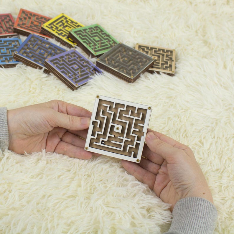 Hands holding a small wooden labyrinth white maze with a metal bead, in front of a colorful assortment of similar maze maze in various colors like red, blue, green, yellow, and more. All displayed on a soft cream fur background.