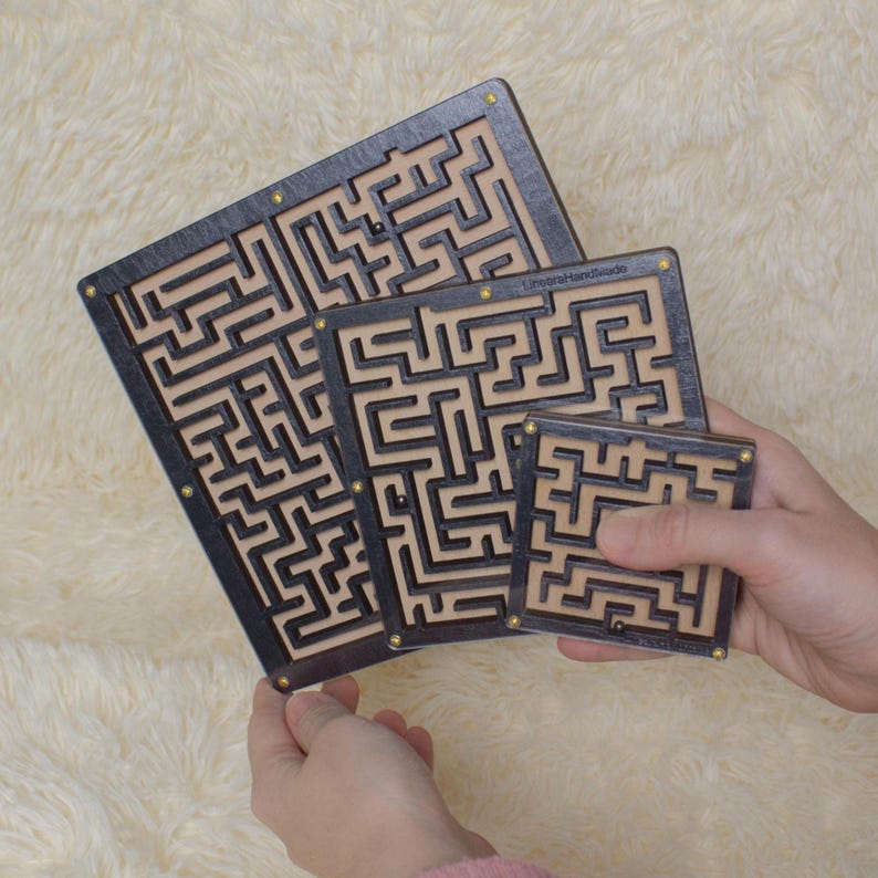Hands holding three black plywood labyrinth mazes in small, medium, and large sizes against a soft beige background, showcasing a handmade maze game set.