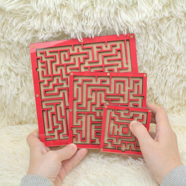 Three sizes of red wooden labyrinth mazes held in hands, each featuring a unique maze design and a one metal bead. Displayed against a soft, fluffy cream background.