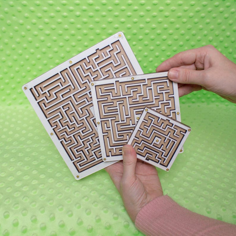Hands holding three white plywood labyrinth mazes in small, medium, and large sizes against a soft green background, showcasing a handmade maze game set.