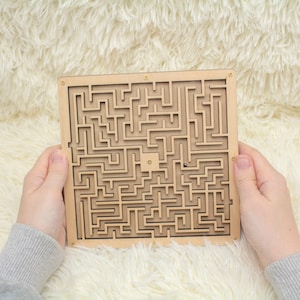 May include: A wooden maze game with a small metal ball inside. The maze is made of light brown wood and has a complex design. The ball is visible in the centre of the maze.