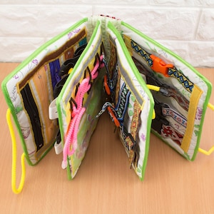 Peut inclure: Un livre d'activités sensoriel ouvert, avec une bordure verte, présentant diverses textures et attaches. Il comprend des fermetures éclair, des boucles et des lacets roses, jaunes et bleus. Le livre est conçu pour l'exploration tactile.