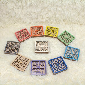 Ten small wooden marble maze toys displayed on a soft cream background, each labeled with a different color option: Red, Orange, Yellow, Green, Blue, Dark Blue, Purple, Black, White, and Without Dyes. The image shows the variety of available color choices for the maze toy.