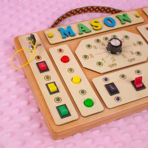 May include: Wooden activity board with colorful buttons, switches, and a dial. The name "MASON" is spelled out in blue, green, and yellow letters. Features a key and a patterned handle. A fun and engaging toy for children.