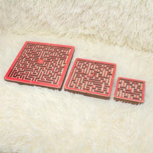 May include: Three wooden maze puzzles of varying sizes. Each puzzle has a red frame and a complex maze design. The mazes are set against a white, fluffy background.