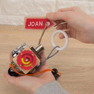 Peut inclure: Un jouet en bois avec une spirale rouge et jaune, un engrenage argenté et un anneau blanc attaché à un cordon gris. Le jouet est étiqueté avec une étiquette en tissu rouge qui dit "JOAN".