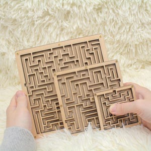 May include: Three wooden maze puzzles of different sizes. The puzzles are made of light-coloured wood with a complex maze design carved into the surface. The puzzles are held against a soft, white background.