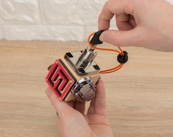 Può includere: Un cubo di legno con un design rosso e bianco e un buco della serratura in metallo. Il cubo è tenuto in una mano e una corda nera e arancione è attaccata al cubo.