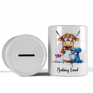 May include: White ceramic money box with a slot on top. The front of the box features a cartoon illustration of a brown Highland cow wearing an apron and holding a spatula in front of a blue stand mixer. The text "Baking fund" is printed below the illustration.