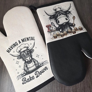 May include: A pair of oven gloves featuring a cartoon cow chef design. One glove has the text "Having a Mental Bake Down". The other has a black textured grip and a cow chef with cupcakes. The gloves are made of cream-colored fabric.