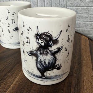 May include: White ceramic money bank featuring a black and white illustration of a Highland cow wearing headphones and dancing. Musical notes are scattered around the cow. The bank has a slot on top for inserting money.