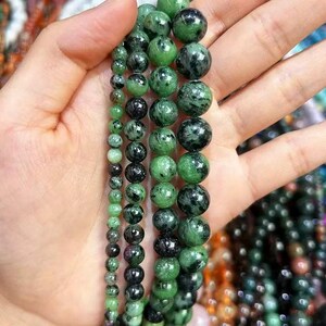 May include: Four strands of round, polished gemstone beads in varying sizes. The beads feature a mottled pattern of deep forest green, black, and occasional flecks of dark pink, arranged in vertical rows from smallest to largest diameter.