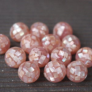 Könnte beinhalten: Eine Gruppe von rosa und weißen Muschelperlen mit einem Mosaikmuster. Die Perlen sind rund und haben eine leicht irisierende Oberfläche.