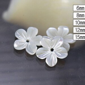 May include: Three white, flower-shaped beads with a pearlescent sheen. Each bead has five petals and a central hole. Sizes are listed as 6mm, 8mm, 10mm, 12mm, and 15mm. The beads are on a dark surface.