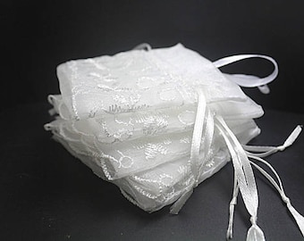 Puede incluir: Un conjunto de bolsas blancas, transparentes, de encaje con cordón y con delicados bordados. Las bolsas están atadas con una cinta de satén blanca.