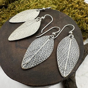 May include: Silver leaf-shaped earrings with detailed veining, displayed on a dark wooden surface. The earrings have a hook closure and a textured finish, resembling real leaves. The background features green moss.