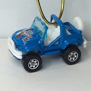 May include: A blue toy jeep with black wheels, a white bumper, and a clear windshield. The jeep has a white spare tire and a gold-colored loop for hanging. The jeep has the text "Rugo" and the number "28" on the hood.