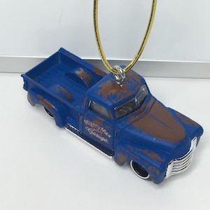 May include: A blue toy truck ornament with a distressed, rusty appearance. The truck has the words "Mighty Max Garage" printed on the side. A gold-colored cord is attached for hanging.