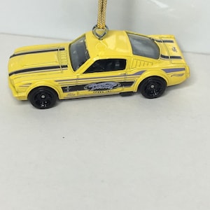 May include: A yellow die-cast model car with black racing stripes and black wheels. The car has the text "Detroit Speed, Inc." on the side and is suspended by a gold string, suggesting it's an ornament. The Ford logo is visible.