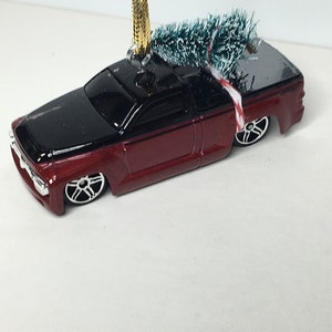 May include: A miniature red and black pickup truck ornament with a small green Christmas tree in the truck bed. The truck has silver wheels and a gold hanging string. A red and white candy cane is tied to the tree.