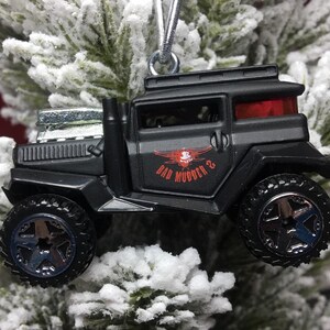 May include: Black toy truck ornament with the text "BAD MUDDER 2" and a skull graphic. The truck has large wheels and a silver engine. The ornament is hanging from a silver string and is set against a snowy, green background.