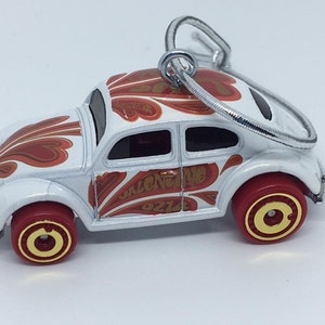 May include: White toy car with red and gold accents. The car features a decorative design on the roof and sides, with the word "VALENTINE" and "OZ14" visible. The wheels are red with gold centers. A silver loop is attached for hanging.