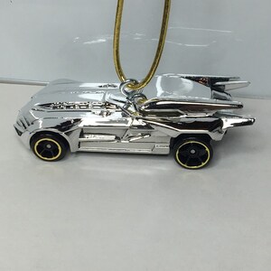 May include: A silver-colored toy car pendant with black and gold wheels, suspended from a gold-colored chain. The car has a sleek, aerodynamic design with fin-like details on the back. The pendant is likely made of metal.