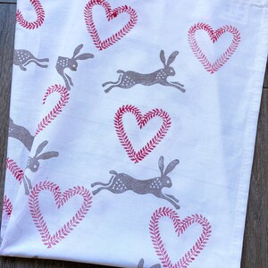 May include: White tea towel with a repeating pattern of red hearts with a leafy border and grey running hares.