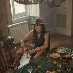 Op de afbeelding: Een vrouw met een hoed en laarzen met luipaardprint zit aan een tafel met een groen tafelkleed. Er staan glazen wijn, een fles champagne en een kom met fruit op tafel.